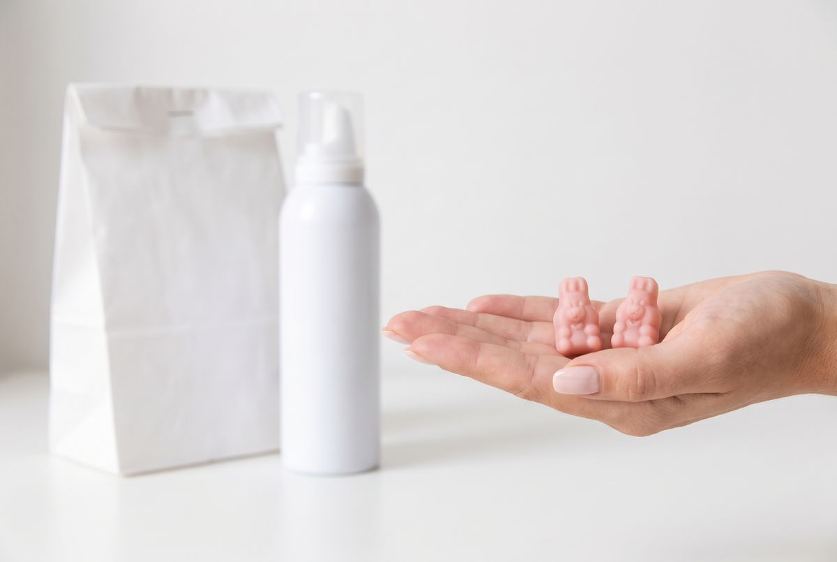Two pink bunny-shaped gummies in an open palm, a pharmacy bag and foam canister behind