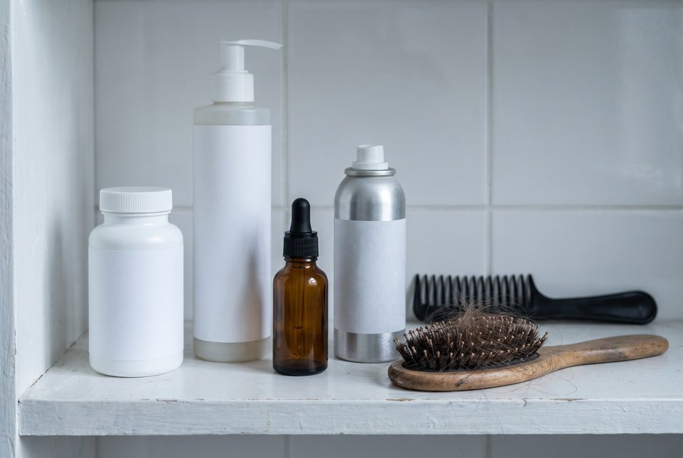 A bathroom shelf of shampoo, oil, foam and a vitamin bottle next to a brush full of hair